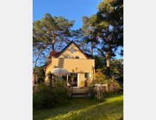 Ein zweigeschossiges Einfamilienhaus mit gelber Fassade und rotem Satteldach, umgeben von hohen Kiefern. Im Vordergrund ein Garten mit Rasen, Pflanzen und einer Terrasse mit Sonnenschirm.