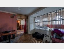 Raum mit hellem Holzboden und einer vertikalen Gittertrennwand, durch die ein weiterer Bereich mit hohen Rundbogenfenstern sichtbar ist. Im Vordergrund stehen vereinzelt Tische, Stühle und gestapelte Gegenstände, während ein offener Durchgang zu einer Treppe mit Holzgeländer führt.