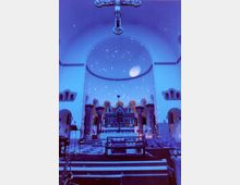 Innenraum einer Kirche mit beleuchteter blauer Atmosphäre. Im Zentrum befinden sich ein kunstvoller Altar und eine Apsis mit runder Kuppel, während ein Kreuz von der Decke hängt; auf dem Boden sind Instrumente und technische Ausrüstung zu sehen.