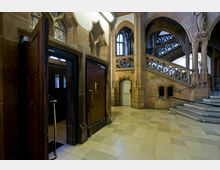 Ein historisches Treppenhaus mit Sandsteinwänden, Spitzbögen und kunstvoll verzierten Balustraden. Der Bereich umfasst geöffnete Holztüren, eine kleine weiße Tür mit Spitzbogenrahmen und eine breite Treppe mit verzierten Geländern.