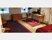 Arbeitszimmer mit hellem Boden, einem langen Holztisch mit Computer und Drucker, einem Stuhl sowie zwei farbigen Teppichen. Im Raum befinden sich eine Holztruhe, Lampen, orangefarbene Stühle und ein Sofa mit roter Decke.