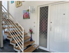 Eingangsbereich eines Hauses mit einer weißen Tür und einem seitlichen Milchglasfenster. Links daneben führt eine Treppe mit Holztufen und weißem Geländer nach oben, an der Wand hängen bunte Bilder, und eine Vase mit rosa Lilien steht auf einem Treppenabsatz.