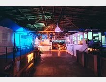 Industriehalle mit sichtbarer Holzbalkendecke, bunten Lichteffekten in Blau und Orange sowie mehreren kleinen Räumen mit Glasfenstern auf erhöhten Plattformen. Im Hintergrund befinden sich weitere Gebäudestrukturen und industrielle Elemente.