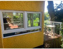 Gelbes Haus mit strukturiertem Putz, links ein Fenster mit Blick in einen hellen Innenraum. Rechts angrenzend ist eine kleine Holzterrasse mit Geländer, Tisch und Stuhl, umgeben von Bäumen und viel Grün.