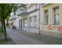 Straßenansicht eines Altbaus mit verputzter Fassade und dekorativen Elementen über dem Eingang. Auf einer angrenzenden Fassade sind rote Graffiti zu sehen, während der Gehweg mit Betonplatten und Pflastersteinen gestaltet ist; ein Baum und geparkte Motorroller sind im Vordergrund erkennbar.