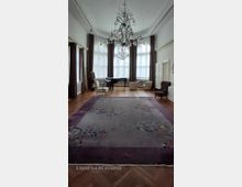 Großer, heller Salon mit hohen Decken, zentralem schmiedeeisernen Kronleuchter und umlaufenden hohen Fenstern mit dunklen Vorhängen. Auf Parkettboden liegt ein großer gemusterter Teppich; an den Wänden stehen ein Flügel und mehrere Sessel.