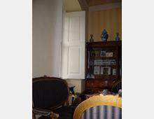 Ein historisch eingerichteter Raum mit verzierten Stühlen, einem Holzschrank mit eingerahmten Bildern und Dekorationsgegenständen, darunter blaue Porzellanvasen. Eine weiße, offene Fensterklappe lässt Tageslicht in den Raum.