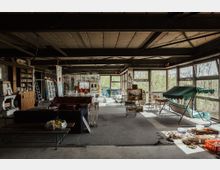 Ein großer, offener Raum mit industriellem Charakter, geprägt von freiliegenden Balken und großen Fenstern, die viel Licht hereinlassen. Der Raum ist mit unterschiedlichen Möbelstücken wie Sofas, Tischen und einer Schaukel ausgestattet, umgeben von Gemälden, Bücherregalen und einigen verstreuten Werkzeugen; Graffiti bedecken stellenweise die Wände.