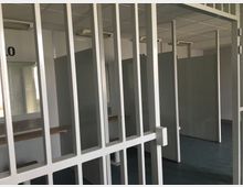 Blick durch ein weißes Metallgitter in einen Innenraum mit grauen Trennwänden und mehreren nummerierten Kabinen, beleuchtet von Deckenleuchten. Links sind ein Fenster und eine Bank zu sehen, der Boden ist dunkelblau.