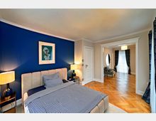 Schlafzimmer mit einer dunkelblau gestrichenen Wand, einem beige gepolsterten Bett mit gestreifter Bettwäsche und Nachttischen mit Lampen. Im Hintergrund ein angrenzender Raum mit Parkettboden, großen Fenstern mit blauen Vorhängen und einem Sessel unter einer Hängelampe.