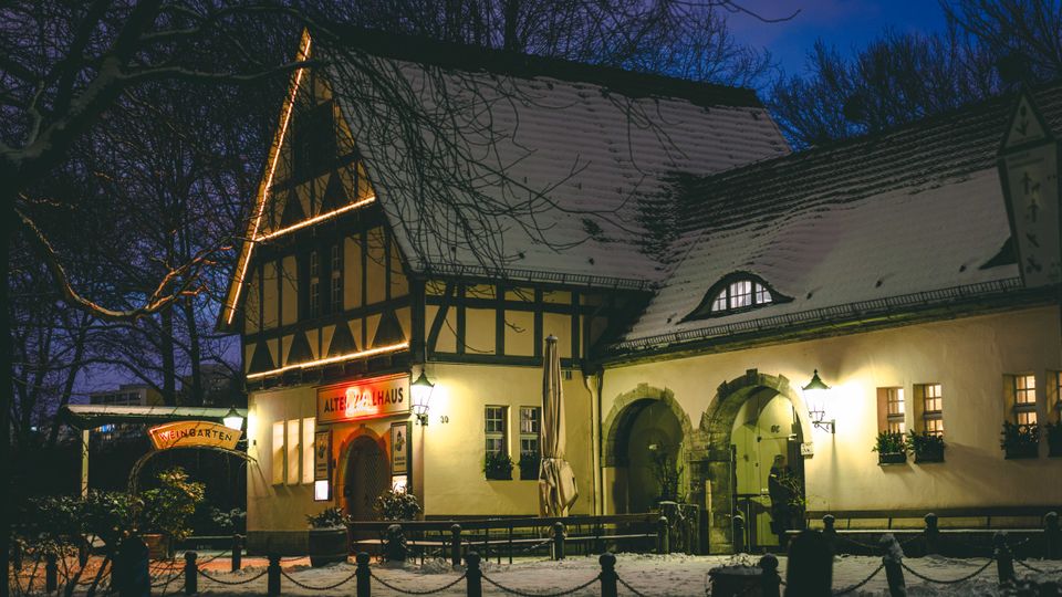 Nächtliche Außenansicht eines historischen Gasthauses im Fachwerkstil mit schneebedecktem Satteldach, beleuchteten Fensterrahmen und einem Eingangsschild „Altes Zollhaus“. Vor dem Gebäude liegt ein verschneiter Hof mit Bänken, Kettenpfosten und kahlen Bäumen im Hintergrund.