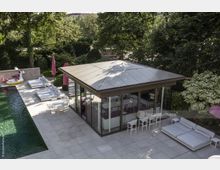 Modernes Gartenhaus mit Glaswänden und Flachdach auf einer großen Terrasse, umgeben von Bäumen und Grünflächen. Neben dem Haus befinden sich ein Pool, Sonnenschirme, weiße Liegen und ein aufblasbares Schwimmtier.