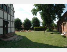 Grüner Hof mit Rasenfläche, umgeben von einem Fachwerkhaus auf der linken Seite, einer roten Backsteinmauer rechts und mehreren Bäumen, darunter gepflegte Hecken und blühende Beete im Hintergrund. Eine weiße Bank steht am Rand der Wiese nahe der Mauer.