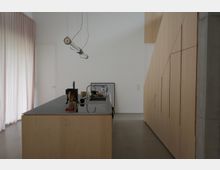 Minimalistische Küche mit einer Holzinsel und einer Arbeitsplatte aus dunklem Material, darauf stehen Küchenutensilien und Geschirr. Rechts befindet sich eine Holzverkleidung mit vertikalen Linien, links ein großer Vorhang vor einem Fenster.