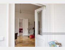 Innenansicht eines Altbaus mit hoher Decke und weiß gestrichenen Wänden. Zu sehen sind geöffnete zweiflügelige Türen, Parkettboden, ein grauer Sessel, ein roter Hocker sowie ein Bereich mit Sofa und Bildern im Hintergrund.