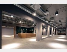 Modernes Innenraumdesign mit Betonboden, sichtbarer Deckenkonstruktion und freiliegenden Lüftungsrohren. Im Vordergrund befindet sich eine Holztheke vor einer Wand mit dunkelgrünen Fliesen, umgeben von offenen Durchgängen und Spotbeleuchtung.