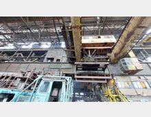 Innenansicht einer alten Industriehalle mit rostigen Stahlträgern, einer großen gelben Laufkatze mit Haken und einer verglasten Fahrerkabine. Im Vordergrund ist ein veralteter, blau lackierter Maschinenwagen zu sehen, und im Hintergrund dominiert die Stahlkonstruktion der Halle.