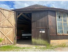 Holz- und Backsteingebäude mit einer großen, geöffneten Holztür, die den Blick in einen Abstellraum mit Fahrzeugen freigibt. Rechts befinden sich alte Glasfenster mit teilweise beschädigten Scheiben, darüber ein Dach mit Moosbewuchs, umgeben von einer ländlichen Umgebung.