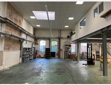 Industriehalle mit grüner Betonbodenfläche, einer Kombination aus unverputztem Mauerwerk und weiß gestrichenen Wänden, sowie sichtbaren Rohren an der linken Wand. Rechts sind moderne Stahlpodeste mit Büros oder Lagerräumen, und im Hintergrund sind Fenster und ein Tor zu sehen, die Tageslicht hereinlassen.