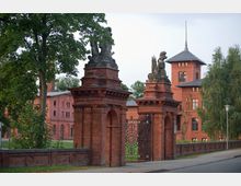 Historisches Backsteinportal mit zwei steinernen Figuren und verzierten Eisentoren, umgeben von alten roten Backsteingebäuden und Bäumen, im Hintergrund ein Turm mit spitzem Dach.