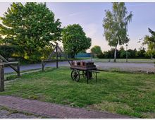 Eine grüne Wiese mit einem hölzernen Handkarren, der Ziegelsteine transportiert, steht im Vordergrund. Im Hintergrund befinden sich Bäume, ein gepflasterter Gehweg, eine Straßenlaterne sowie eine ruhige Straße.