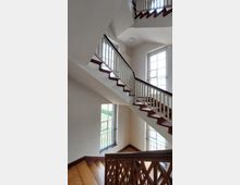 Treppenhaus mit hölzernem Geländer und Treppenstufen, weiß gestrichenen Wänden und großen Fenstern mit Gitterstäben, die Tageslicht hereinlassen und den Blick auf eine grüne Außenanlage ermöglichen.