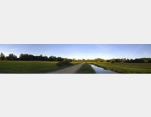 Panorama einer ländlichen Landschaft mit einer unbefestigten Straße, die durch grüne Wiesen führt. Rechts der Straße verläuft ein schmaler Wassergraben, und im Hintergrund sind Bäume an der Waldrandgrenze zu sehen, eingerahmt von einem blauen Himmel.