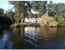 Ein weißes Gebäude mit schwarzen Fensterrahmen steht am Ufer eines Sees, umgeben von großen Bäumen und Rasenflächen. Das ruhige Wasser des Sees spiegelt die Vegetation und das Haus wider.