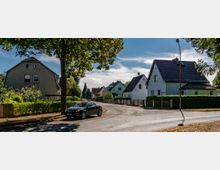 Straßenszene in einem Wohngebiet mit Einfamilienhäusern in traditioneller Bauweise. Im Vordergrund steht ein geparktes Auto, umgeben von Bäumen, Zäunen und Gärten; im Hintergrund sind weitere Häuser und ein klarer Himmel sichtbar.