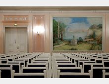 Ein klassisch gestalteter Saal mit Reihen weißer Stühle und einer großen Wandmalerei, die eine Landschaft mit Bäumen, einer Säule, einem Fluss und Bergen im Hintergrund zeigt. Die Wände sind in warmen Tönen gehalten, mit dekorativen Ornamenten über einer Doppeltür und an den Seitenlampen.