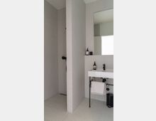 Minimalistisches Badezimmer mit einer offenen Dusche links, die durch eine Mauer abgetrennt ist. Rechts befindet sich ein Waschbecken mit Spiegel, darunter ein Handtuchhalter und ein kleiner Abfalleimer.