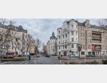 Straßenszene in einer Stadt mit mehrstöckigen Wohngebäuden im historistischen Stil, charakterisiert durch verputzte Fassaden, Erker und verzierte Dachformen. Im Vordergrund eine Kreuzung mit Verkehrszeichen, Fußgängern, Radfahrern und geparkten Autos, umgeben von kahlen Bäumen.