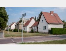 Eine ruhige Wohnstraße mit Einfamilienhäusern mit weißem Putz und roten Satteldächern, umgeben von kleinen Gärten und Hecken. Straßenschilder zeigen die Kreuzung von "Glärnischweg" und "Mariendorfer Damm", links verläuft ein Gehweg entlang einer Baumreihe.