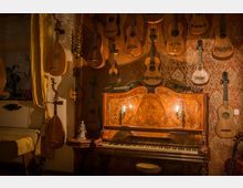 Ein rustikales Zimmer mit roter, gemusterter Tapete, in dem zahlreiche Gitarren, Lauten und andere Saiteninstrumente an der Wand hängen. Im Vordergrund steht ein antikes Klavier aus Holz mit verzierten Details, darüber ein wandmontierter Kerzenhalter mit brennenden Kerzen.