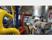 Industrieller Innenraum einer Maschinenhalle mit großen Rohrleitungen in Gelb und Rot, metallischen Ventilen und Tanks. Die Halle hat eine Wellblechdecke, und im Hintergrund sind weitere Rohrsysteme und Anlagen sichtbar.