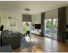 Modernes Wohnzimmer mit hellem Holzboden, grauem Ecksofa und einem runden Glastisch auf einem Teppich. Große Fenster mit Blick auf einen Garten lassen viel Tageslicht herein, ergänzt durch helle Vorhänge und minimalistische Dekoration.