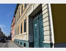 Straßenszene mit einer Reihe von Mehrfamilienhäusern in klassischer Architektur. Die Fassade eines Gebäudes in gelblichen und grünen Tönen mit großen Holz-Doppeltüren ist im Vordergrund zu sehen, daneben ein Gehweg und parkende Autos.