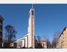 Moderne Kirche mit einem markanten, hohen Glockenturm aus hellem Beton und Backstein, gekrönt von einem Kreuz. Das Kirchengebäude verfügt über große, rechteckige Fenster und steht in einer städtischen Umgebung mit Wohnhäusern und kahlen Bäumen.