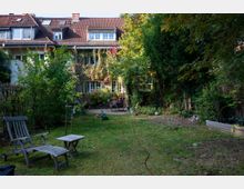 Großer Garten hinter einem mehrstöckigen Wohnhaus mit rotem Ziegeldach und vielen Fenstern, teils von Kletterpflanzen bewachsen. Auf der Rasenfläche stehen Gartenliegen und ein kleiner Tisch; dichte Bäume und Sträucher säumen die Grundstücksgrenzen.