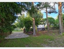 Gartenbereich mit Rasen, Bäumen und einem gepflasterten Weg, der zu einem Holzzaun führt. Im Hintergrund ist ein weißer Kleintransporter neben dem Zaun zu sehen.