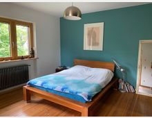 Schlafzimmer mit einem Holzbett, das eine weiße Decke und eine blaue Bettwäsche hat. Die Wände sind teils türkis gestrichen, und ein großes Fenster mit Holzrahmen bietet Blick ins Grüne; daneben befinden sich eine Heizung und Regalflächen mit Büchern und Dekorationen.