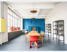 Großer, heller Besprechungsraum mit einer langen Holztafel und Holzstühlen, einer roten Stuhlakzentuierung im Vordergrund. Raum geprägt durch blaue Wand, retro-stilistische Glasvitrinen, große Fenster mit viel Tageslicht und dekorative Elemente wie ein "Grand Hotel"-Schild.