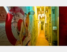 Schmaler, beleuchteter Gang mit rauen Betonwänden, buntem Graffiti, und Wandlampen mit runden Kugelschirmen. Rechts befinden sich nummerierte Türen mit der Aufschrift "Zelle" sowie sichtbare Lüftungsrohre an der Decke.