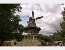 Historische Windmühle mit hölzernen Flügeln und einem steinernen Sockel, umgeben von Bäumen und kleinen Gebäuden, unter einer teils bewölkten Himmel. Die Mühle ist auf einem erhöhten Hügel mit einer steinernen Balustrade errichtet.