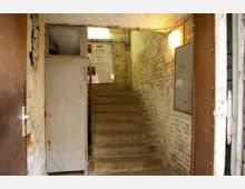 Ein schmaler, teils renovierungsbedürftiger Treppenaufgang in einem Gebäude mit hellen Backsteinwänden und brüchigem Putz. Eine metallene Tür steht offen, während an der Wand ein Plakat mit Informationen angebracht ist und Tageslicht durch einen oberen Bereich eindringt.