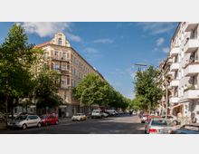 Straßenszene in einer urbanen Umgebung mit mehrstöckigen Wohngebäuden auf beiden Seiten. Links ein historisches Eckgebäude mit dekorativer Fassade und Jahrzahl &quot;1896&quot; auf dem Giebel, rechts ein moderneres Wohngebäude. Entlang der Straße stehen geparkte Autos, und Bäume säumen den Gehweg.