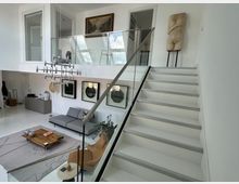 Helles, modernes Wohnzimmer mit offener Galerie, einer weißen Treppe mit Glasgeländer, einer grauen Couch, einem flachen Holztisch und dekorativer Kunst an den Wänden. Große Fenster und minimalistische Einrichtung prägen den Raum.