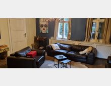 Wohnzimmer mit einer braunen Ledercouch und einem passenden Sessel, modernen Wandfarben (eine Wand in Blau), einem großen Fenster mit beigefarbenen Vorhängen sowie verschiedenen dekorativen Elementen wie einem Pop-Art-Wandbild und einem Kronleuchter. Ein kleiner Couchtisch steht in der Mitte auf einem Teppich.