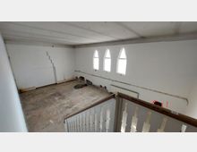 Ein leerer Raum mit weiß gestrichenen Wänden, drei schmalen, spitz zulaufenden Fenstern und sichtbaren Bauschäden an der unteren Wand. Im Vordergrund befindet sich ein Treppengeländer aus Holz mit vertikalen Aussparungen.