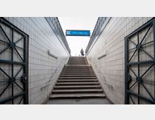 Eine Treppenaufgang einer U-Bahn-Station mit Fließen an den Wänden und Metallgeländern. Oben ist ein blaues Schild mit der Aufschrift "U Uhlandstraße" sichtbar, und eine Person geht die Treppe hinauf.